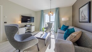 Ein modernes Wohnzimmer mit einem Flachbildfernseher, grau gepolsterten Sessel, einem Couchtisch und einem Sofa mit türkisen und beigen Kissen.