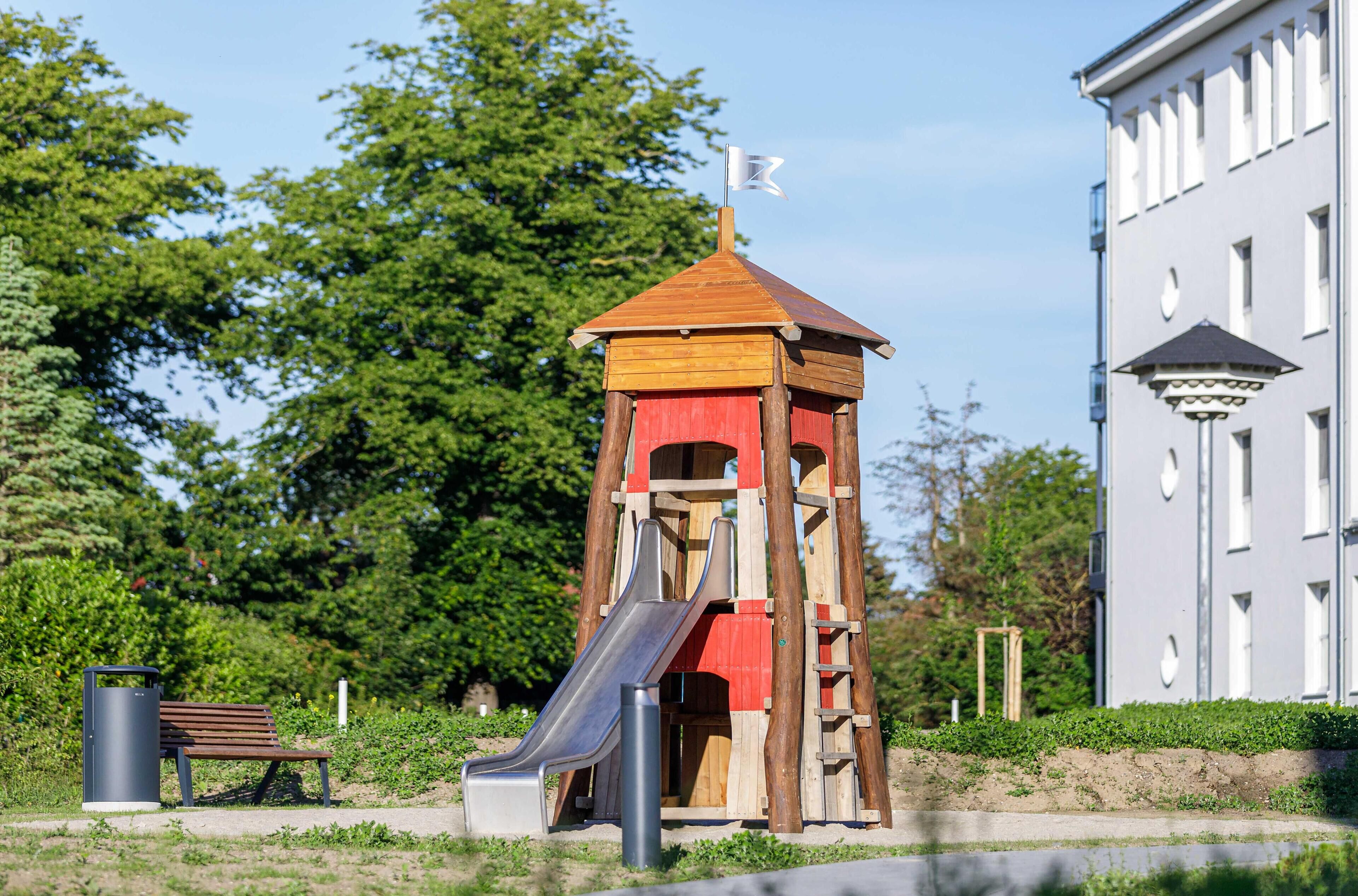 Een speeltuin met een glijbaan en een bankje, omringd door groen en een gebouw.