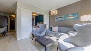 A modern living room with a grey sofa, armchairs, a coffee table, and a dining area. There are two wall-mounted paintings and a hanging light fixture.
