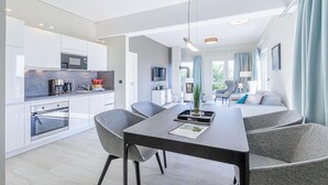 A modern kitchen with built-in appliances, a dining area with grey upholstered chairs, and a view of the outdoors through large windows. - Beautiful 5-star vacation apartment Rundblick between the Bodden and the Baltic Sea (Wustrow)