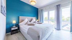 A modern bedroom with a large bed, a nightstand with a lamp, and a view of the outside through large windows. - Beautiful 5-star vacation apartment Rundblick between the Bodden and the Baltic Sea (Wustrow)
