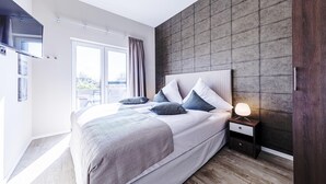 A modern bedroom with a large bed, a nightstand with a lamp, and a view of the outdoors through a sliding door. - Humboldtsuite Wustrow - 5 stars, wellness, balcony & underground parking (Wustrow)