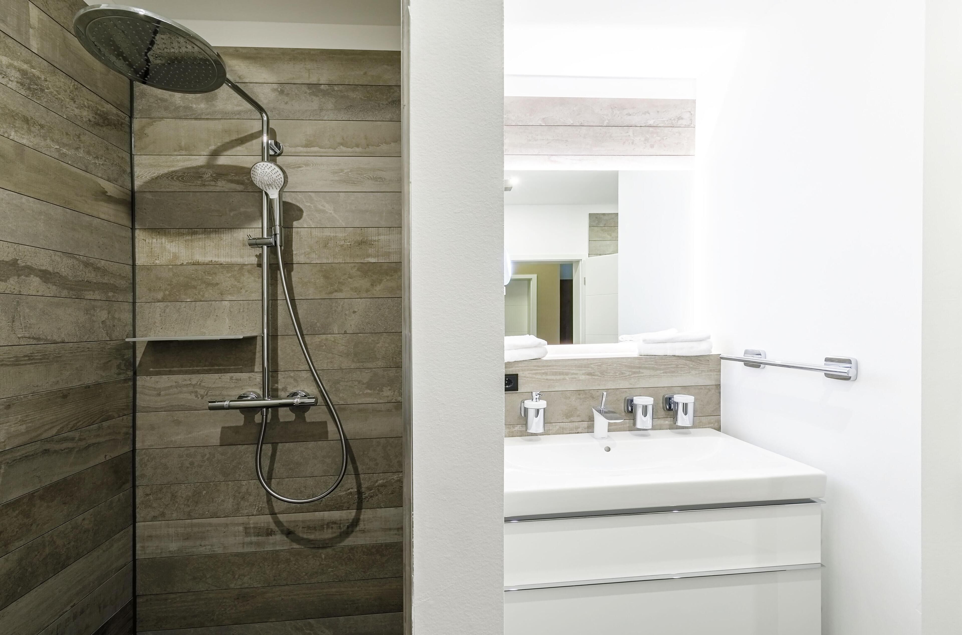 A modern bathroom with a wooden wall, a shower with a handheld showerhead, and a white sink with a mirror above it.