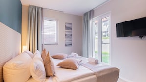 Una cama bien hecha con almohadas y una colcha, una ventana con cortinas, un televisor de pantalla plana montado en la pared y un cuadro en la pared.