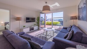 A modern living room with a grey sofa set, a white coffee table, and a TV mounted on the wall. There is a large window providing a view of the outside.