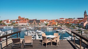 A waterfront terrace with wicker chairs and tables overlooking a marina with boats, historic buildings, and a tower.
