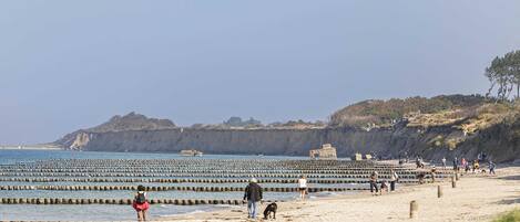 A beach with a rocky cliff in the background, wooden groynes, and people walking along the shore.