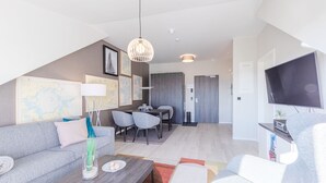 Una sala de estar moderna con un sofá gris, una mesa de centro y un televisor. Hay una zona de comedor con una mesa y sillas, y una cocina al fondo.