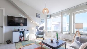 Una sala de estar moderna con un televisor de pantalla plana, una estantería, un sillón gris, una mesa de centro y grandes ventanales con vistas a la ciudad.