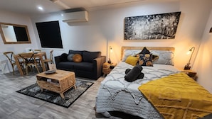 Egy modern hálószoba, amelyben egy nagy ágy, egy kanapé, egy étkezőasztal székekkel és egy mennyezeti ventilátor található.