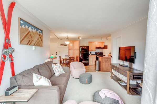 A living room with a sofa, ottoman, and a television. There is a kitchen in the background with cabinets and a refrigerator. - Skiing & Hiking: Mtn-view Condo in Durango! (Durango)