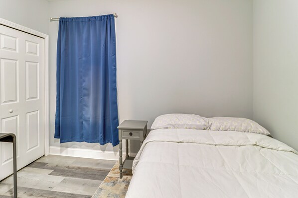Un dormitorio con una cama blanca, una mesita de noche pequeña, una cortina azul y una puerta blanca.