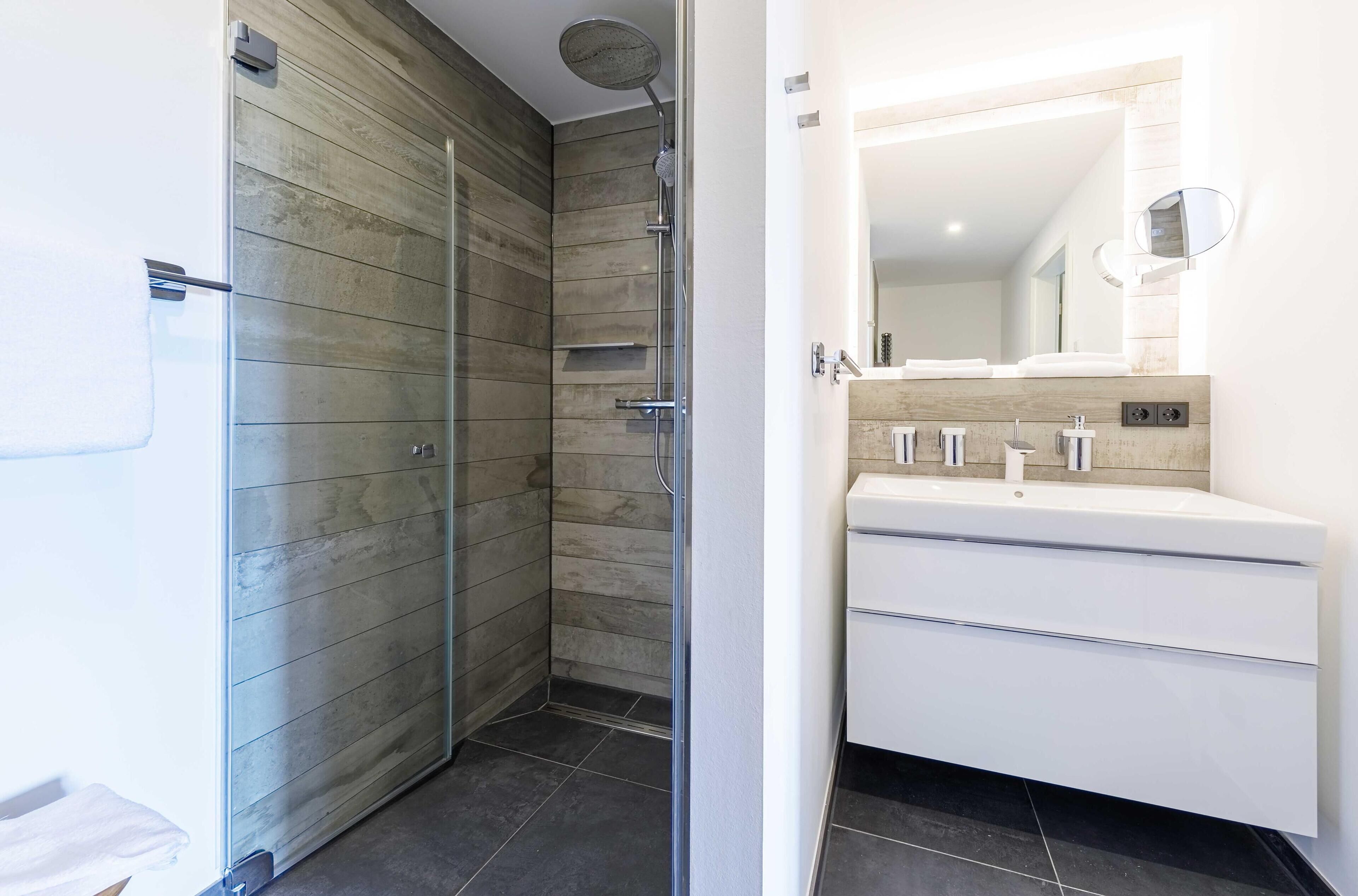 Un baño moderno con ducha envuelta en vidrio, un lavamanos blanco con espejo y suelo de baldosas.