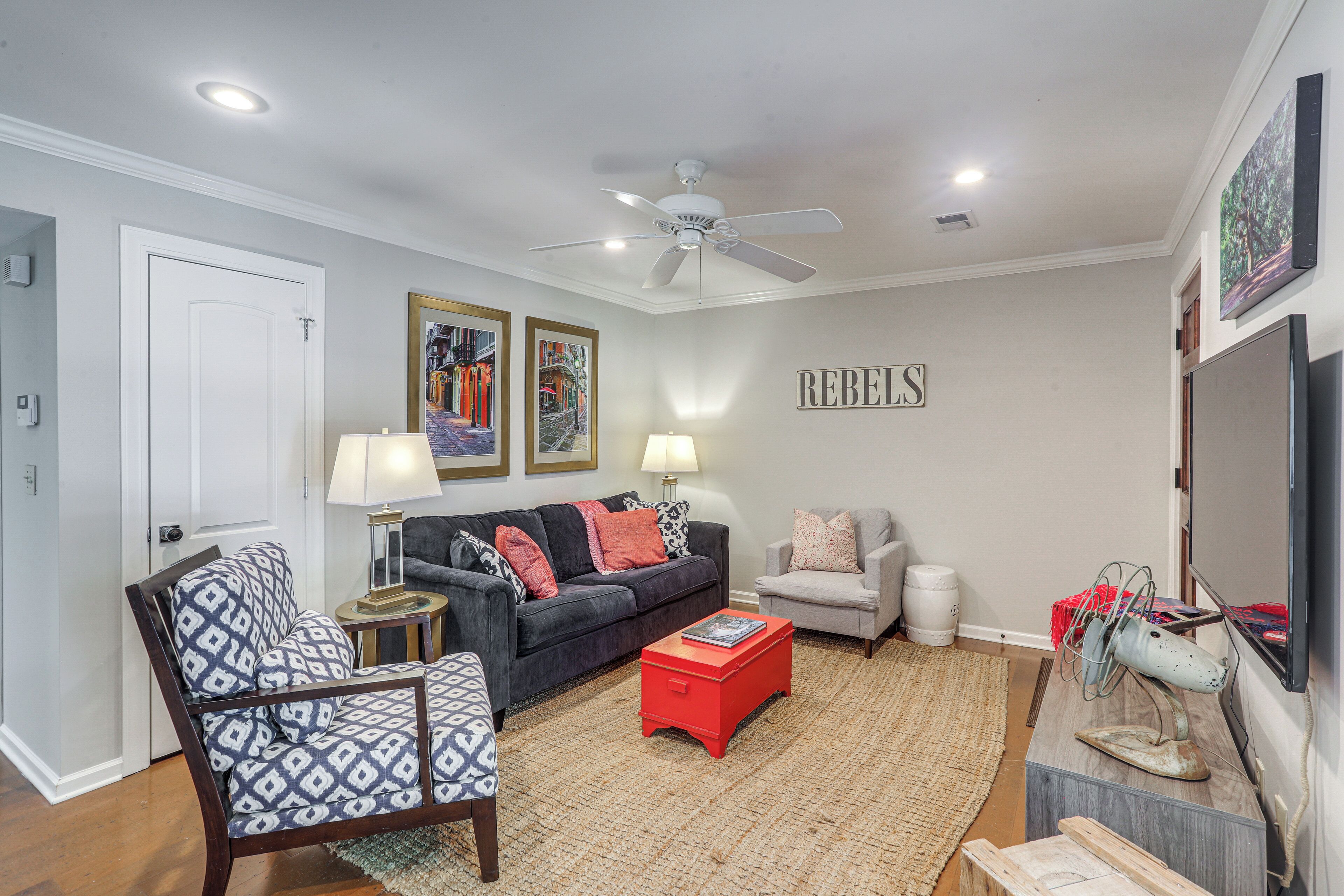 A living room with a dark grey sofa, a patterned armchair, a red ottoman, a ceiling fan, and a wall-mounted TV.