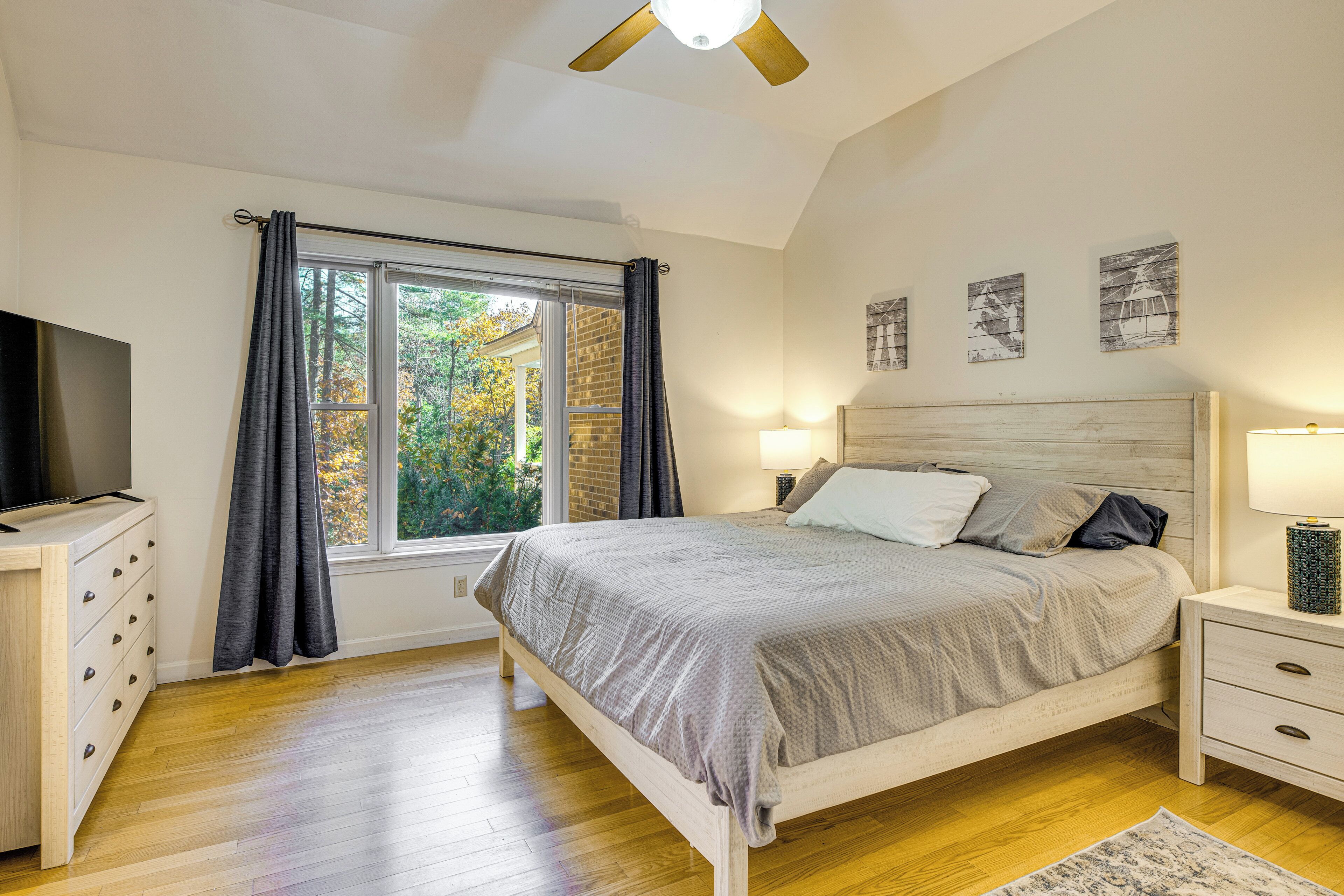 A bedroom with a bed, a nightstand, a TV, a window with curtains, and a ceiling fan.