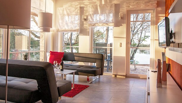 A modern living room with a sofa, a coffee table, and a red rug. There is a large window with a view of trees and a body of water. - Vacation apartment "Fischadler" with terrace in Jabel an der Müritz (Jabel)