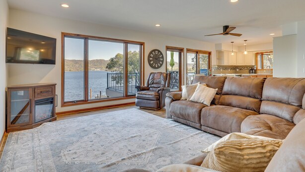 A spacious living room with a large window overlooking a body of water, a leather sofa, a fireplace, and a flat-screen TV mounted on the wall. - Dock & Views: Waterfront Home in Wabasha! (Wabasha)