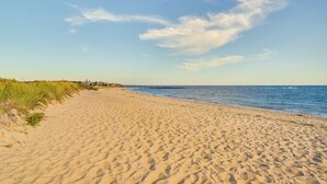 A sandy beach with gentle waves, a clear sky, and distant buildings. - Bike to Private Beach: Cape Cod Escape w/ Sunroom (East Falmouth)