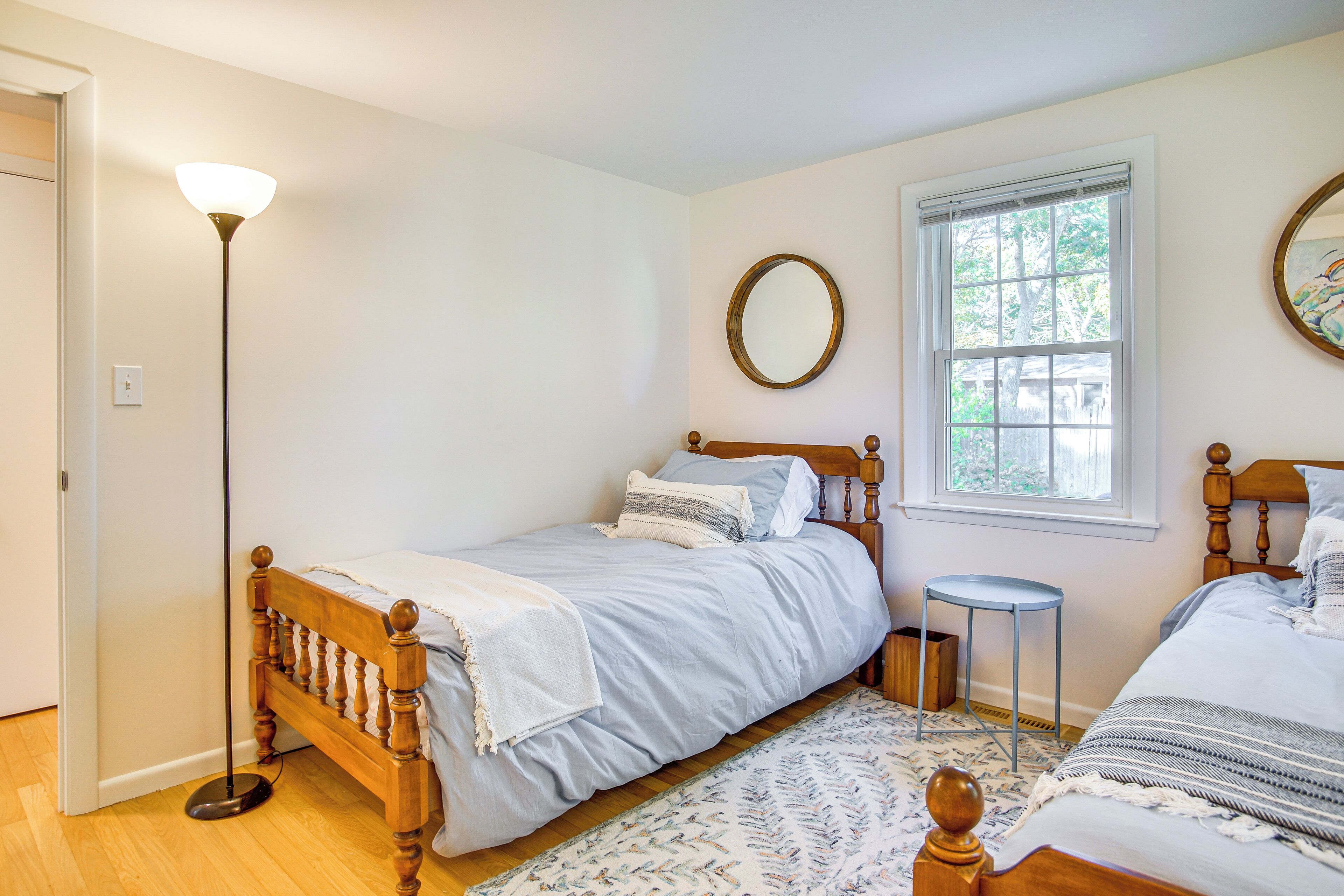 A bedroom with two wooden beds, a standing floor lamp, a small round mirror, and a window with blinds.