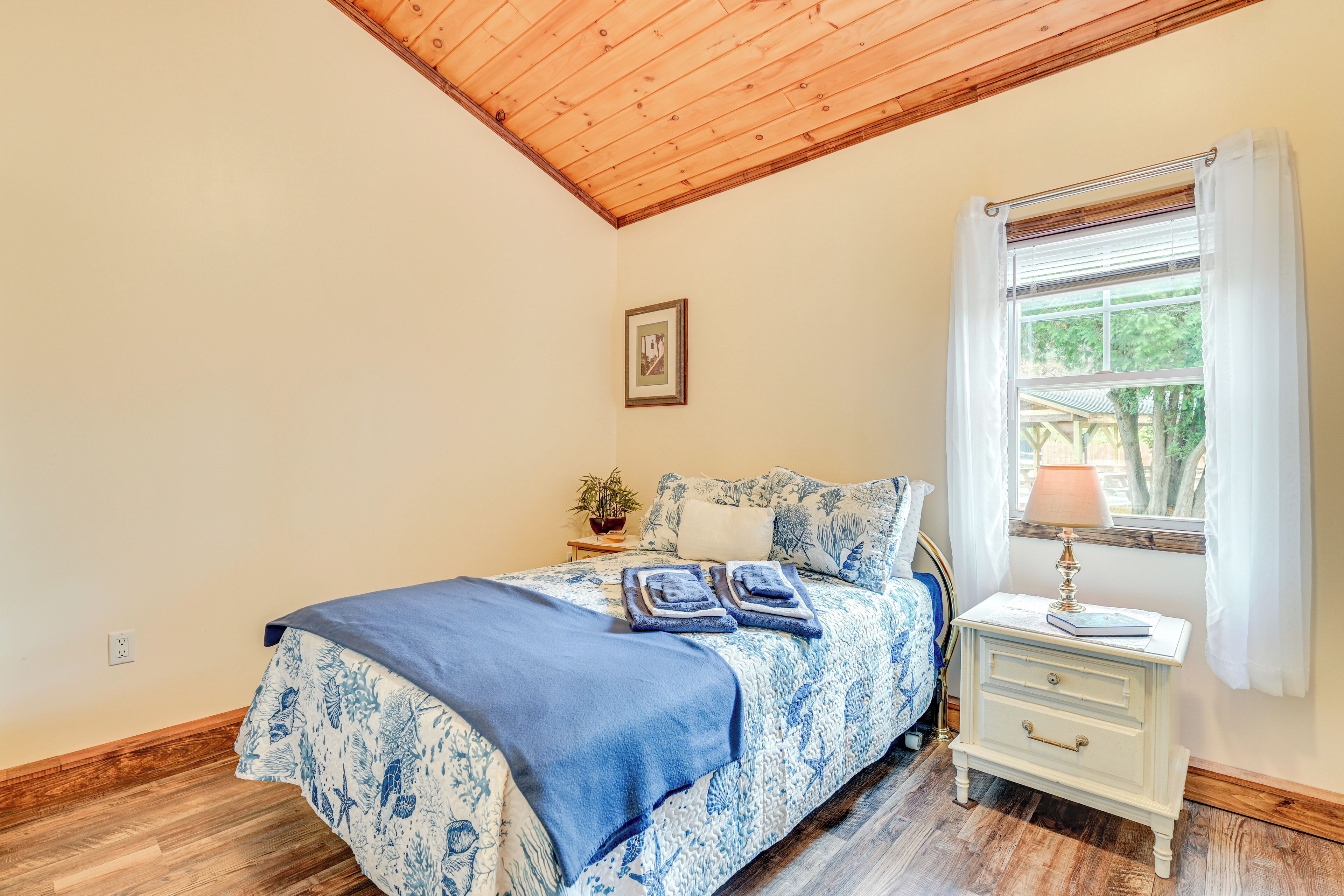 Ein Schlafzimmer mit Holzboden, einem Bett mit blumigem, blauem Überwurf, einem Nachttisch mit Lampe und einem Fenster mit Vorhängen.