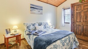A bedroom with a wooden ceiling, a bed with a blue and white floral bedspread, a nightstand with a lamp, and a window with a view of trees. - 2 Mi to Lake Champlain: Cabin w/ River Access! (Peru)