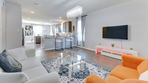 Ein modernes Wohnzimmer mit einer Couch, einem Couchtisch und einem Fernseher. Im Hintergrund ist eine Küchenzeile mit Barhocker und einem Esstisch zu sehen.