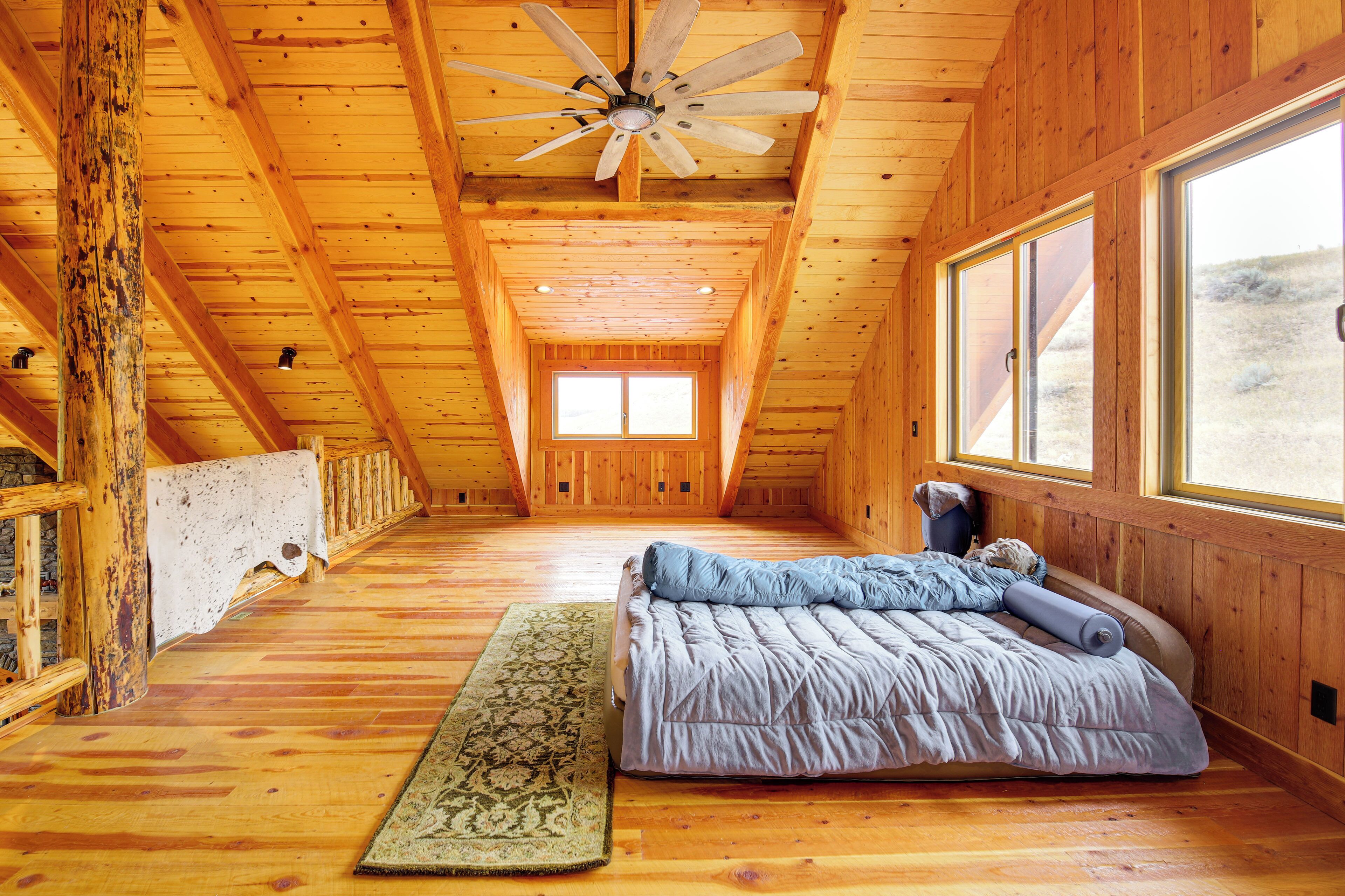 Interior de una cabaña de madera con una cama, un ventilador de techo y ventanas amplias.