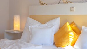 Een opgemaakt bed met witte en gele kussens, een bedlampje en een lichtschakelaar aan de muur.