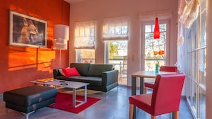 Een moderne woonkamer met een bank, salontafel en een rode fauteuil. Er hangt een schilderij aan de muur en een raam met uitzicht op bomen.