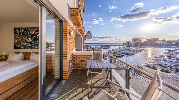 En balkon med trægulv, en sofa og et bord. Der er udsigt over en lystbådehavn med både og en bro i baggrunden.