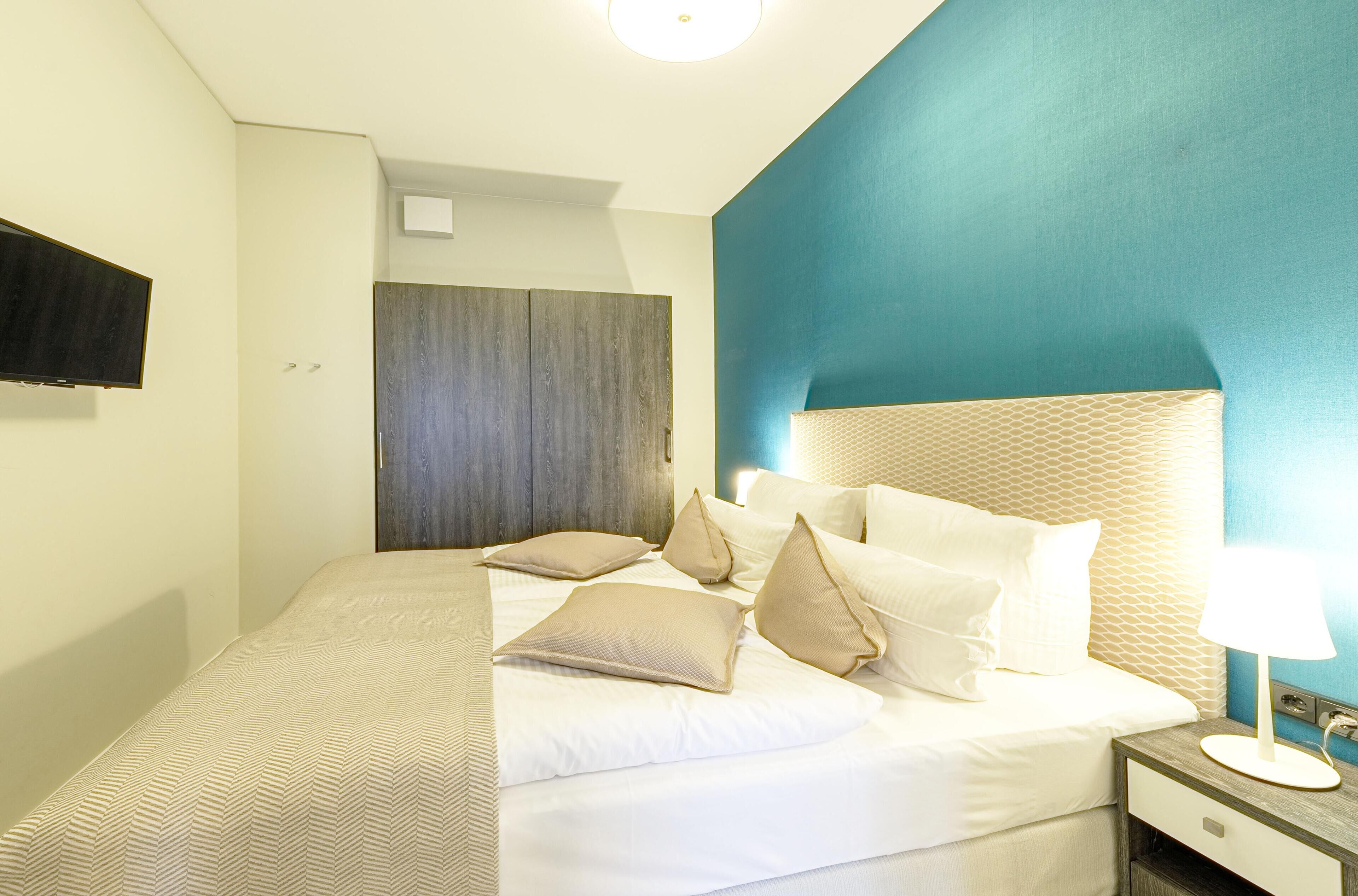 A neatly made bed with multiple pillows, a bedside table with a lamp, and a wall-mounted TV.