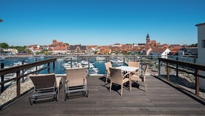 A wooden deck with wicker chairs and a table overlooking a marina with boats and buildings in the background.