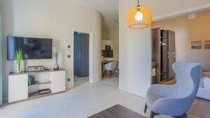 A modern living room with a flat-screen TV, a white entertainment unit, a grey armchair, and a dining area with a table and chairs.