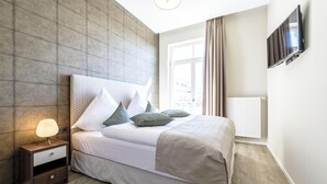 Una cama bien hecha con cabecera, una mesita de noche con lámpara, y un televisor montado en la pared.