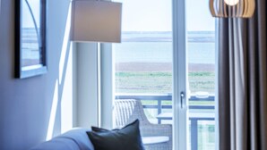 Habitación de hotel con cama, lámpara, vista al mar a través de un ventanal grande y un balcón con una silla.