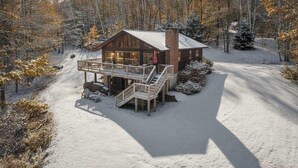 Una cabana de fusta amb terrassa i escala, envoltada d'un paisatge nevós i arbres.