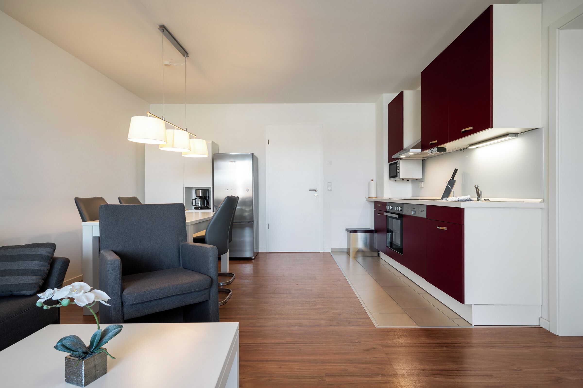 A modern kitchen with red cabinets, a built-in oven, and a microwave. There is a gray armchair and a white table with a vase of flowers.