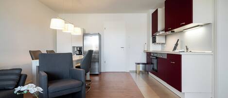 A modern kitchen with red cabinets, a built-in oven, and a microwave. There is a gray armchair and a white table with a vase of flowers.