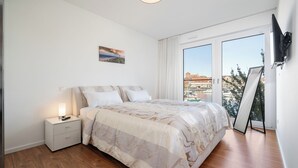 Een slaapkamer met een bed, nachtkastje, televisie en een groot raam met uitzicht op gebouwen.