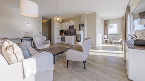 Ein modernes Wohnzimmer mit einer Couch, einem Couchtisch und Sesseln, eine Küche mit Geräten und ein Essbereich mit Tisch und Stühlen.