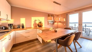 A modern kitchen with a wooden table and chairs, a coffee maker, and a view of the sea through large windows.