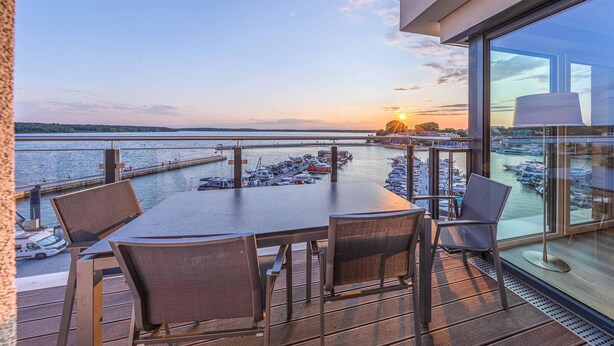 A terrace with a table and chairs overlooking a marina with boats and a sunset view.