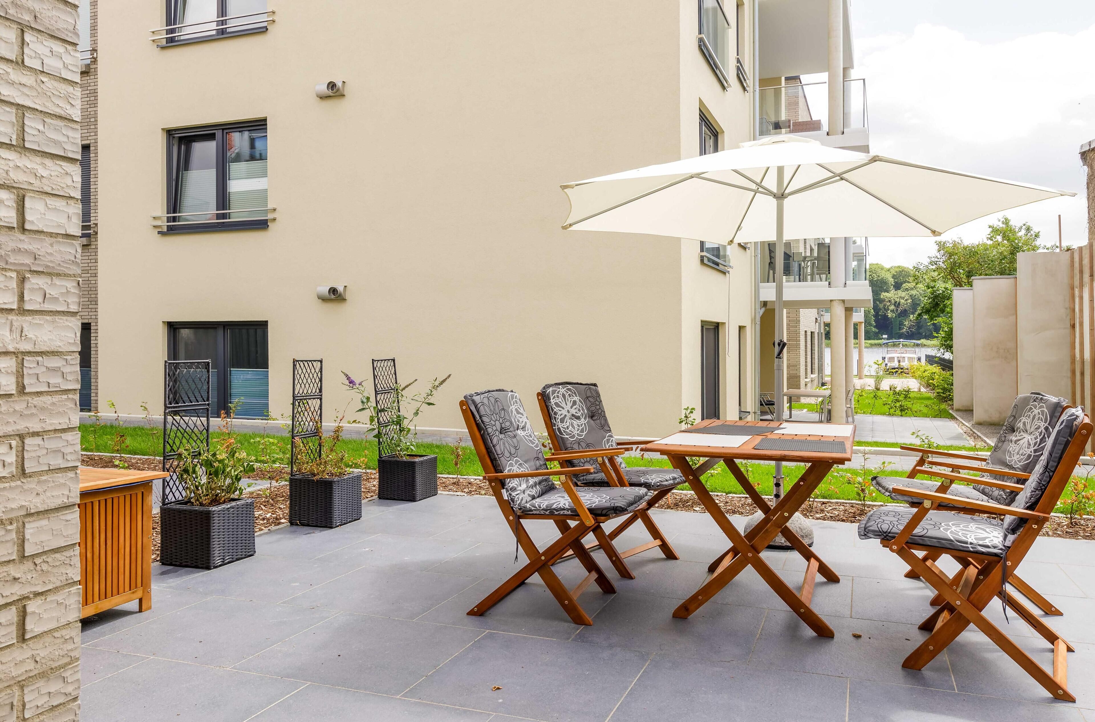 Un patio exterior con una mesa y cuatro sillas, rodeado de plantas en macetas y vista a un edificio.