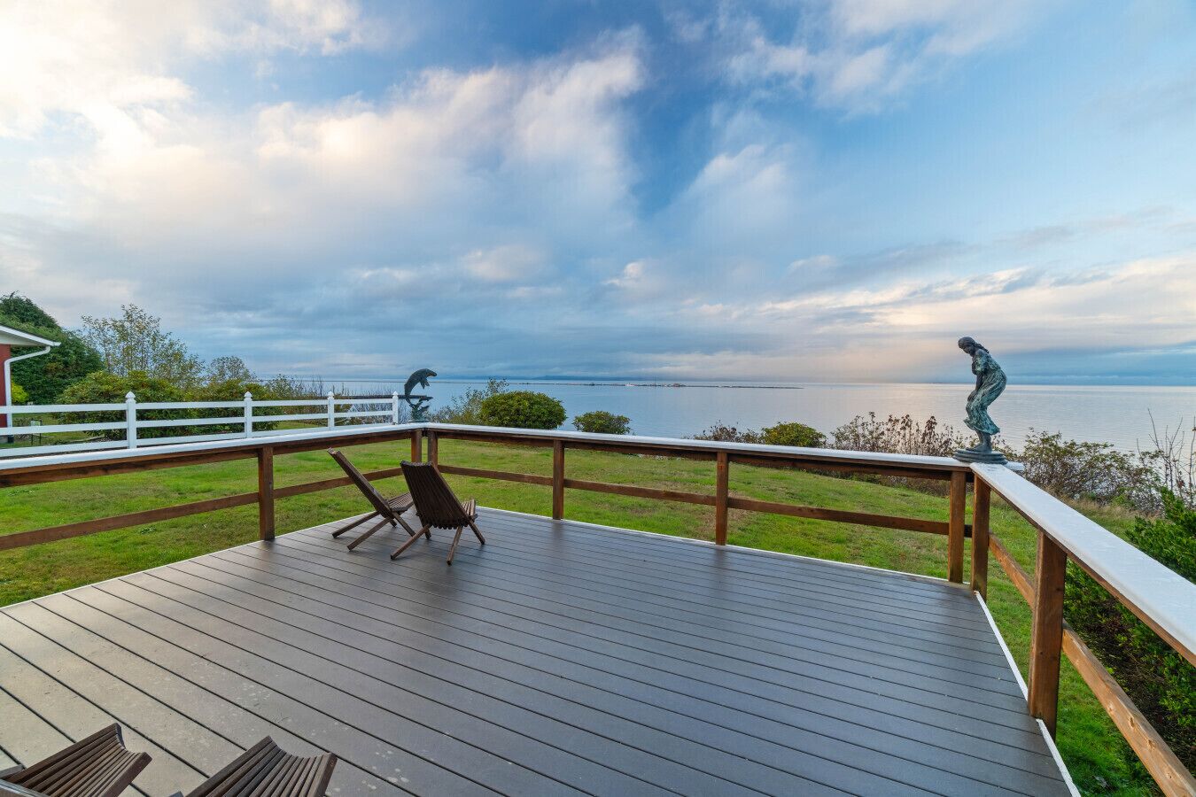 A wooden deck with two lounge chairs, a white fence, and a view of the sea.