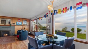 A modern living room with a fireplace, a dining table with chairs, and a view of the sea through large windows. - Water Edge Coast-Guard Cottage-Panorama View- (Port Angeles)