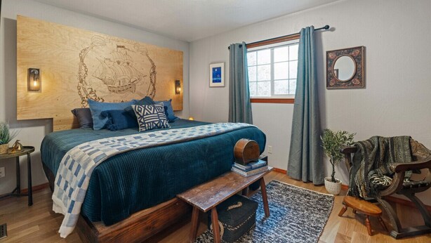 A bedroom with a large bed, a wooden headboard, a nightstand, a bench, a window with curtains, a mirror, and a wall-mounted artwork. - Water Edge Coast-Guard Cottage-Panorama View- (Port Angeles)