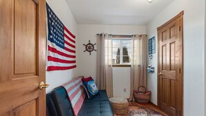 A hallway with a wooden door, a black leather sofa with a flag pillow, a ship's wheel decoration, and a patterned rug. - Water Edge Coast-Guard Cottage-Panorama View- (Port Angeles)