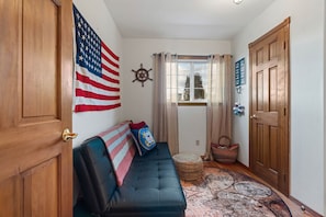 A hallway with a wooden door, a black leather sofa with a flag pillow, a ship's wheel decoration, and a patterned rug. - Water Edge Coast-Guard Cottage-Panorama View- (Port Angeles)