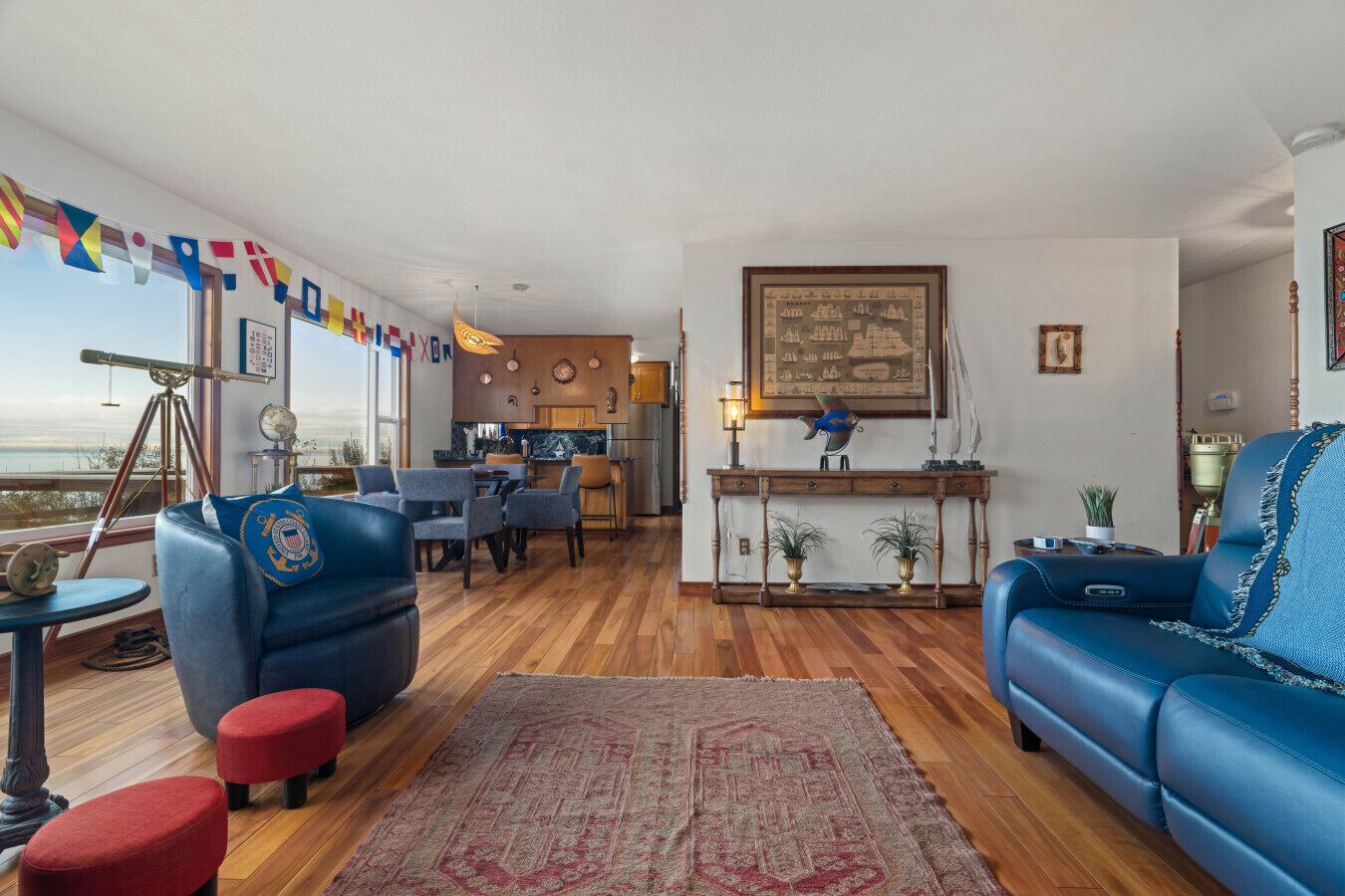 A living room with blue sofas, a dining area, a kitchen, and a view of the sea.
