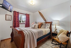 A bedroom with a wooden bed, a nightstand with a lamp, a rocking chair, and a TV mounted on the wall. - Water Edge Coast-Guard Cottage-Panorama View- (Port Angeles)
